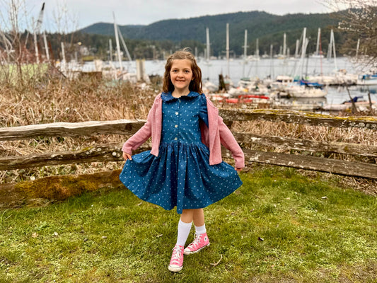 Girl in jean dress & pink sweater smiling in grass wt water behind