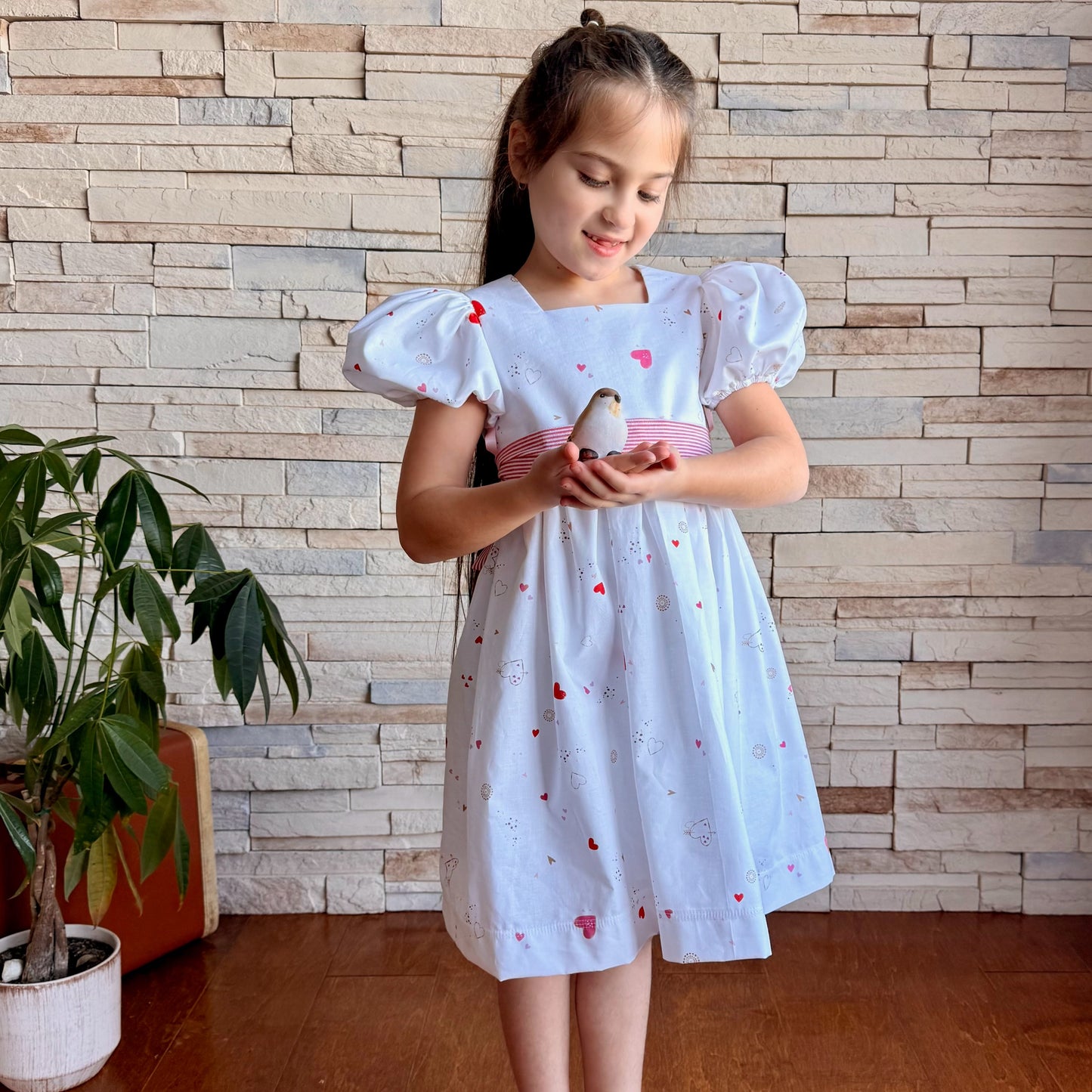 Young girl in a white dress with red hearts holding a small bird against a brick wall.