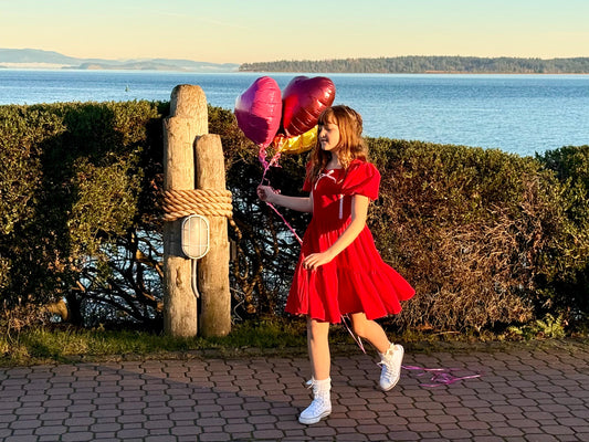 Girl in red dress walking waterside with heart balloons 