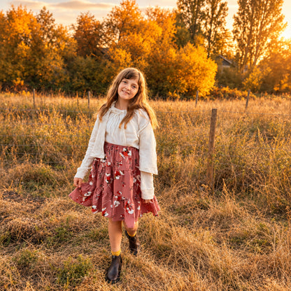 A young girl stands in a grassy field wearing a white long-sleeve blouse and a dusty rose pink skirt with a white floral pattern. The background features dry grass, a wooden fence, and trees with bright yellow autumn foliage.