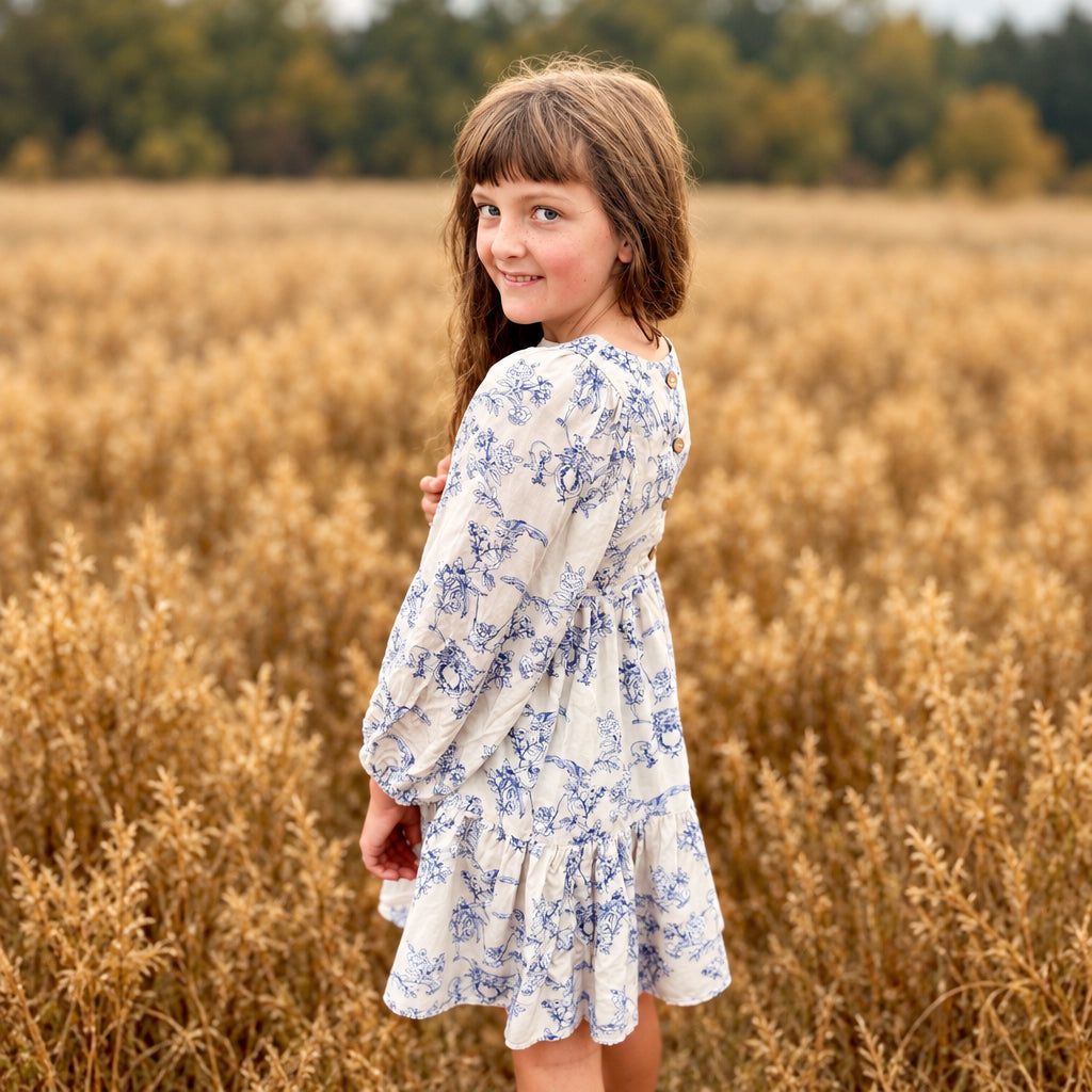 A young girl with brown hair stands in a field of dry golden grass, looking back over her shoulder. She is wearing a long-sleeved cream dress with a blue floral pattern.