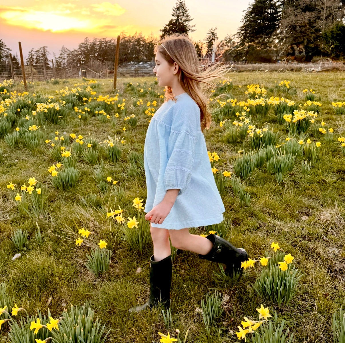 Woman in a light blue dress and black boots standing in a field of yellow flowers with a sunset in the background.