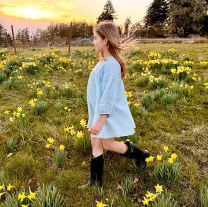 Woman in a light blue dress and black boots standing in a field of yellow flowers with a sunset in the background.