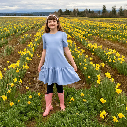 Young girl in a light blue dress and pink boots standing in a field of yellow flowers with a scenic background.