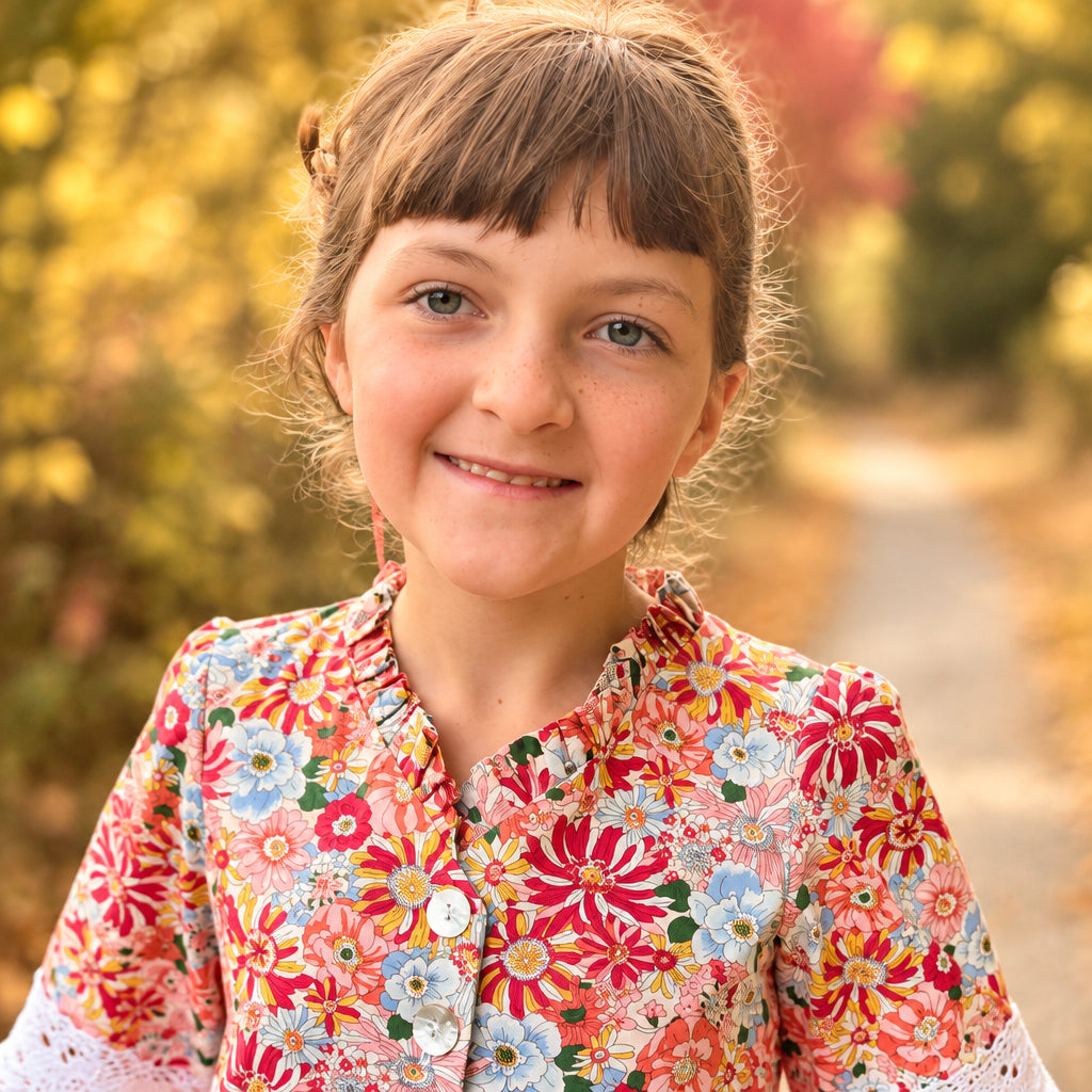 A young girl with brown hair and bangs stands on a dirt path outdoors, wearing a long-sleeve button-front dress with a dense multicolor floral print in red, orange, pink, green, and blue on a light background. The dress has white lace cuffs and a ruffled hem.