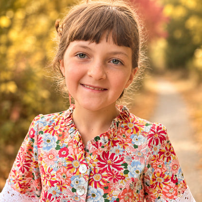 A young girl with brown hair and bangs stands on a dirt path outdoors, wearing a long-sleeve button-front dress with a dense multicolor floral print in red, orange, pink, green, and blue on a light background. The dress has white lace cuffs and a ruffled hem.