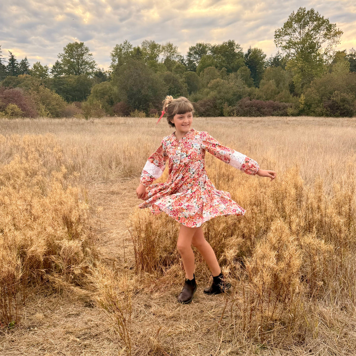 A young girl wearing a red and white floral long-sleeve dress and black boots stands in a field of dry golden grass, holding out her arms as if twirling. Trees and a cloudy sky are visible in the background.