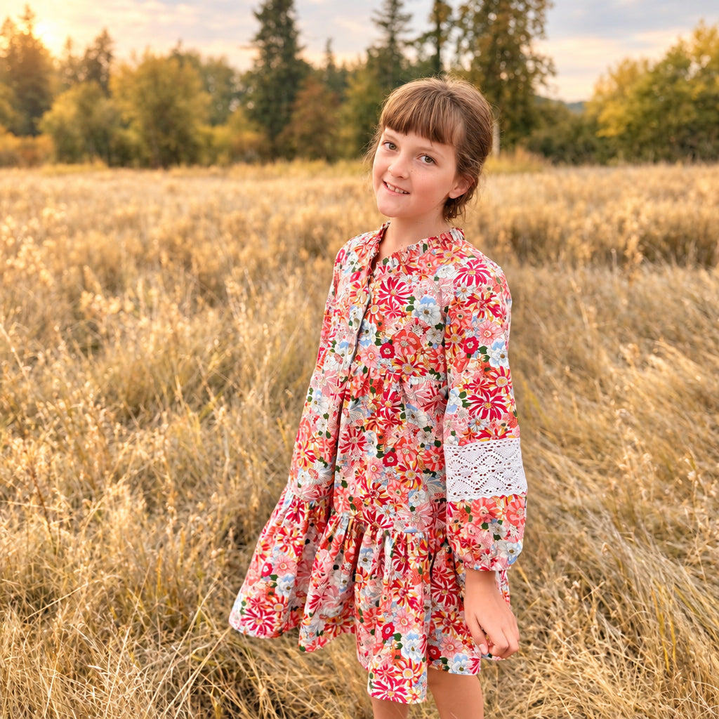 A young girl stands in a dry grassy field wearing a colorful floral long-sleeve dress with white lace cuffs. Trees are visible in the background under a cloudy sky.