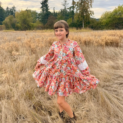 A young girl with brown hair and bangs stands in a field of dry, golden-brown grass, wearing a long-sleeved floral dress with a ruffled hem and black boots.