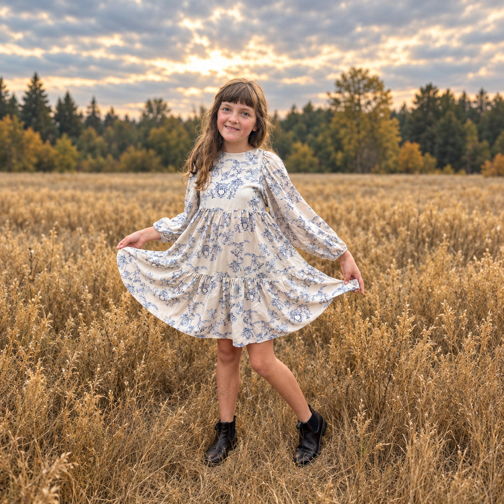 A young girl with brown hair stands in a dry grass field wearing a cream-colored long-sleeve dress with a blue floral pattern and black shoes.