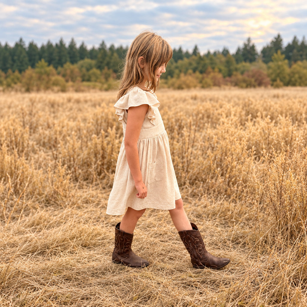 A young girl wearing a cream-colored ruffle-sleeve dress and brown cowboy boots walks through a dry grassy field outdoors.