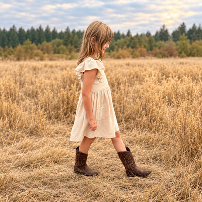 A young girl wearing a cream-colored ruffle-sleeve dress and brown cowboy boots walks through a dry grassy field outdoors.