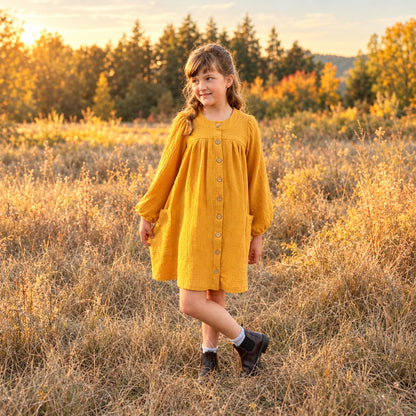 A young girl wearing a mustard yellow long-sleeve button-front dress stands in a grassy field during late afternoon sunlit hours, with trees and blue sky in the background.