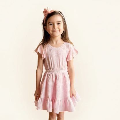 Young girl in a pink dress standing against a brick wall.