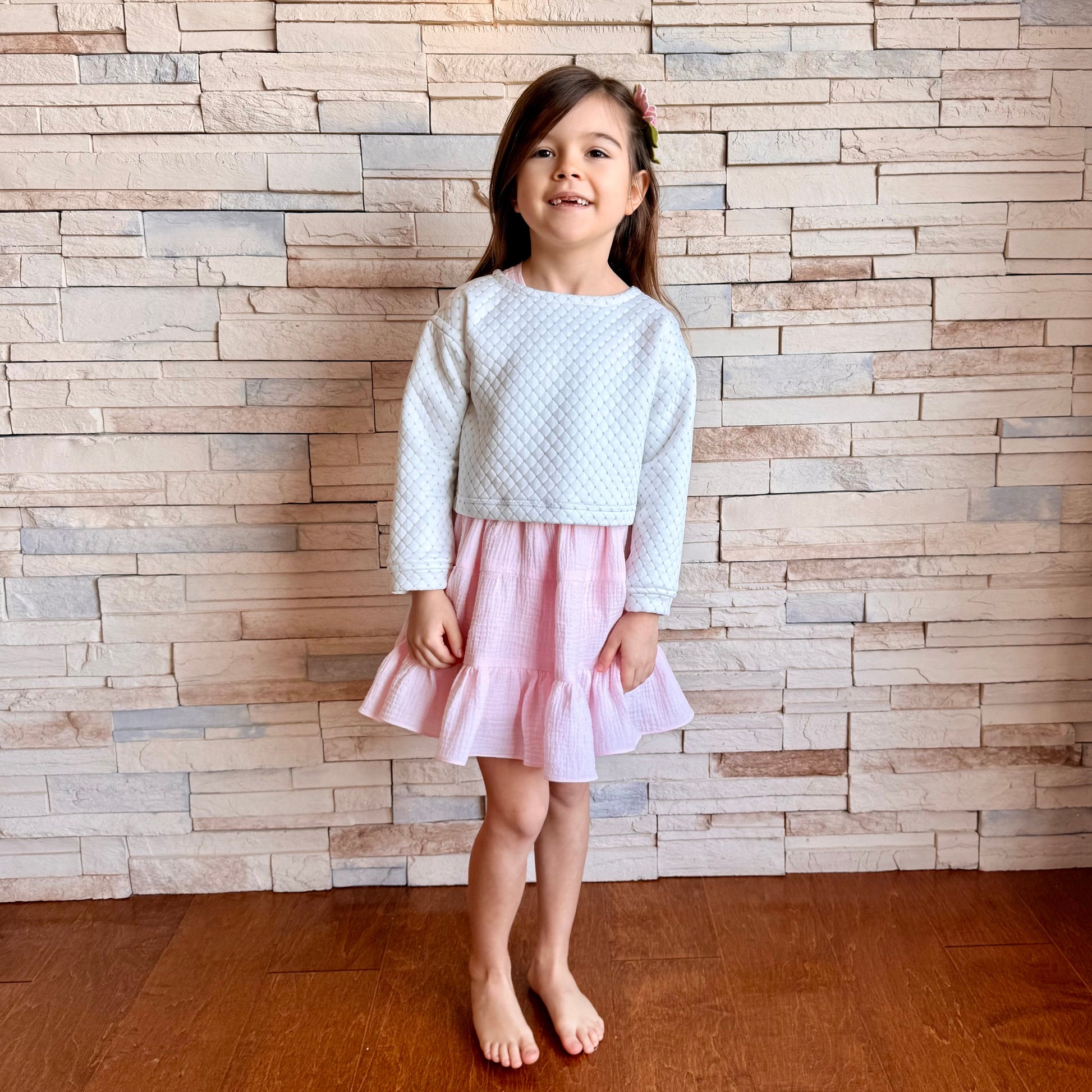 Young girl in a light blue sweater and pink skirt standing against a brick wall.
