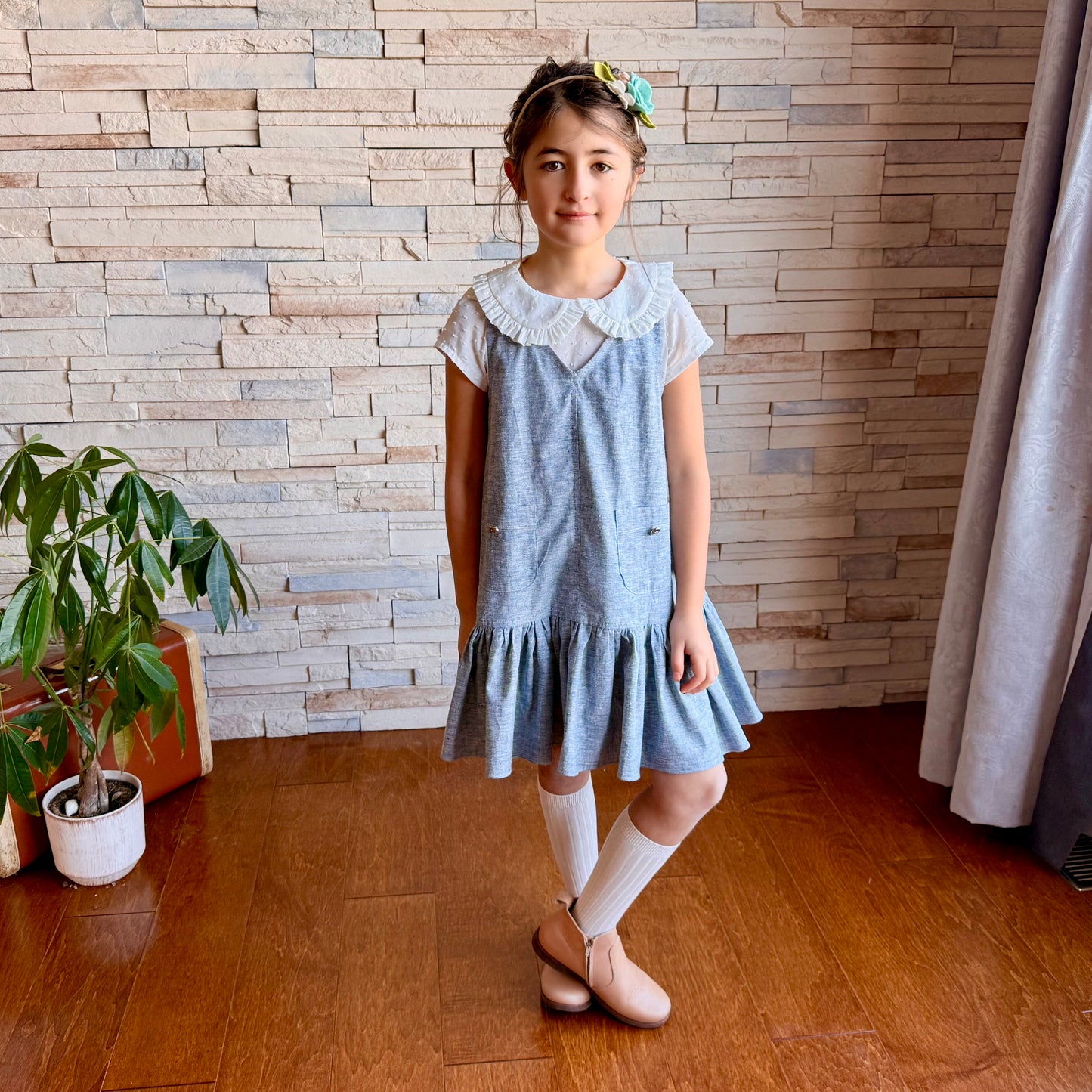 Young girl in a blue dress standing in a room with wooden flooring and a stone wall.