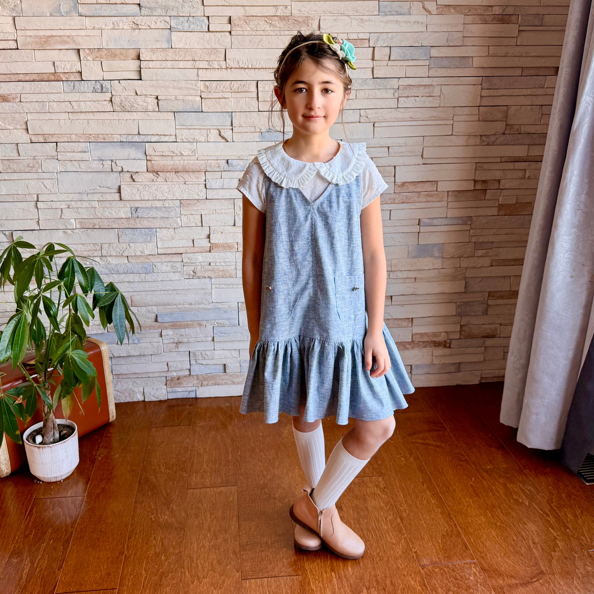 Young girl in a blue dress standing in a room with wooden flooring and a stone wall.
