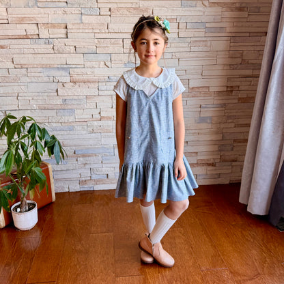 Young girl in a blue dress standing in a room with wooden flooring and a stone wall.