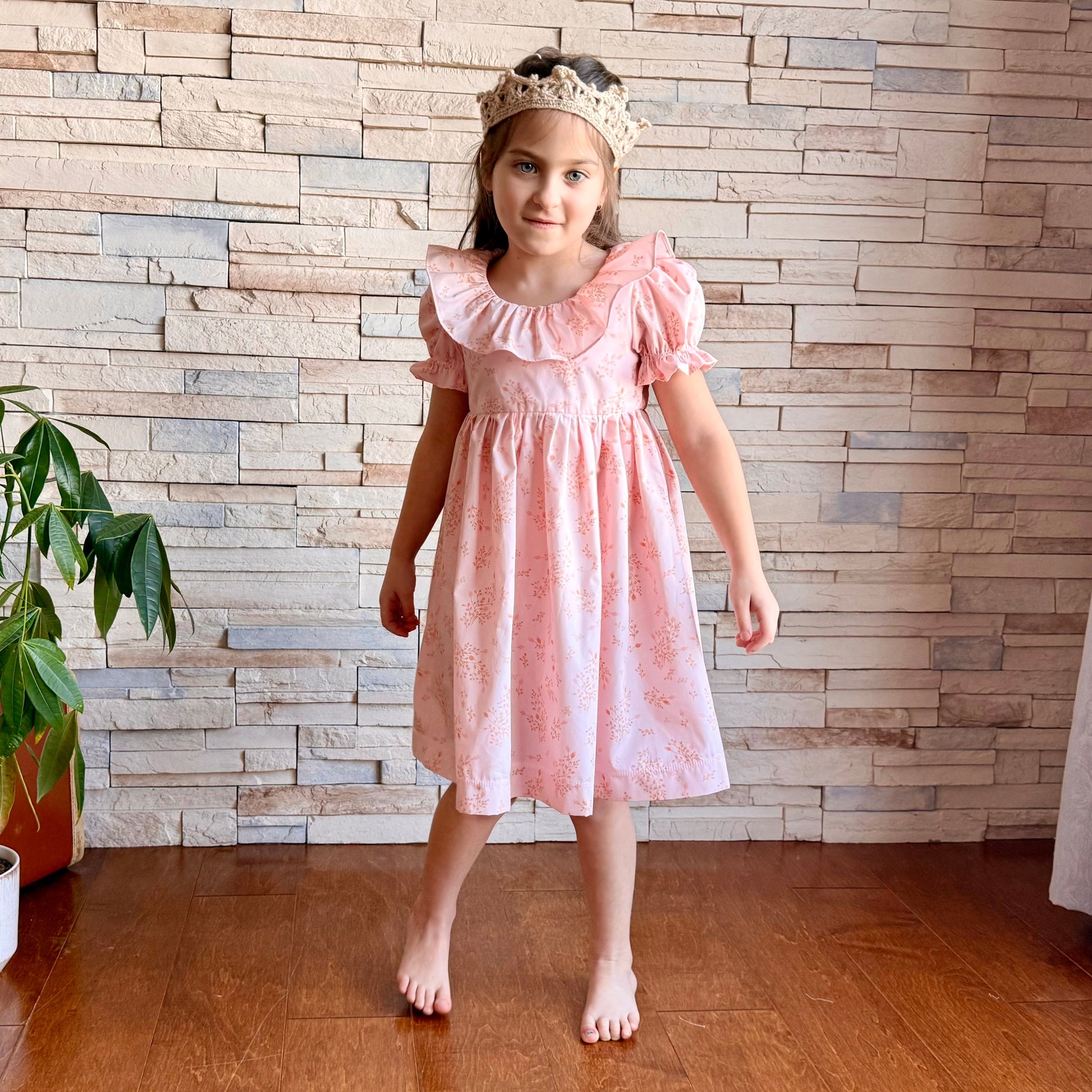 Young girl in a pink dress standing in front of a wooden paneled wall.
