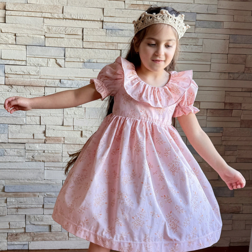 Young girl in a pink dress with ruffled sleeves standing against a stone wall.