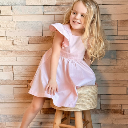 Young girl in a pink dress sitting on a wooden stool against a wooden paneled wall.
