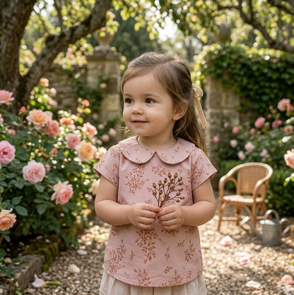 Young girl in a pink floral dress standing in a garden with flowers and a chair in the background.