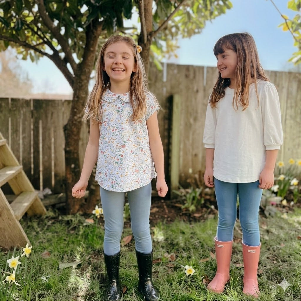 Two young girls standing in a garden with flowers and a wooden fence in the background.